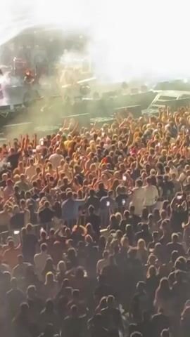 This video shows a large crowd of people at an outdoor concert. The crowd is packed together and many are holding up their phones, likely recording the performance. The stage is visible in the background with bright lights and what appears to be performers. The overall atmosphere is energetic and lively.