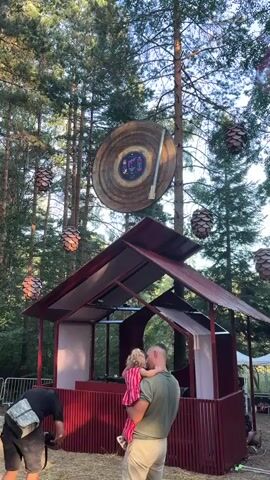 A man holds a young child with blonde curly hair in a pink and red striped dress. The child is facing away from the camera and towards a DJ booth set up under a red, open-sided structure with a sloped roof. The DJ booth contains DJ equipment. Behind the DJ booth, a large, circular object resembling a vinyl record is suspended from a tree by a thin cable. Several cone-shaped decorations hang from trees in the background. Another person, partially visible, stands to the left of the man and child, holding a camera. To the left and in front of the man, a young boy wearing a yellow shirt and green shorts is looking towards the DJ booth. To the right and behind the boy, a young girl with blonde hair, wearing a white top and pink shorts, is looking towards the camera. The scene takes place outdoors in a wooded area with trees and dry grass on the ground.