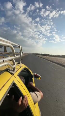 A POV shot from the passenger side of a yellow car driving on a paved road. The passenger's arms are visible out the window, one holding onto the side of the car. The other hand is on the steering wheel. In the distance, the road stretches towards the horizon, flanked by dry, arid land and sparse vegetation. The sky is a vibrant blue with scattered white clouds. The car has a metal rack on its roof. The sun appears to be low in the sky, casting a warm light.