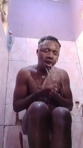 The video shows a shirtless dark-skinned man in a bathroom, sitting on what appears to be a toilet seat. He is wet and water is running down his face and chest. He appears to be washing his face with both hands, and then he starts moving his hands as if he is praying or expressing strong emotion while he vocalizes. The bathroom has pink-tinted tiles on the walls. A tube of toothpaste is affixed to the wall, and there is a faucet and plumbing visible. The lighting appears dim, casting some shadows.
