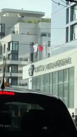 A street-level view from inside a car shows the front of the "Klifton Hotel & Résidences" building. A French flag and another flag wave in the wind on flagpoles in front of the building. A modern-style, white building with a rooftop garden sits beside the hotel. A few other buildings, power lines, and a "Beware of Pedestrians" sign can be seen. The video appears to be taken during the day.