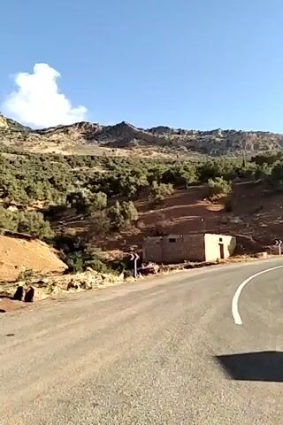 The video shows a rural road in a mountainous region on a sunny day. 

The camera pans from left to right, initially focusing on the road and a small, tan, brick-like structure on the side of the road. The landscape features low-lying, shrub-covered hills.

The camera then pans to capture a road sign with directional arrows, presumably indicating different towns or locations. The text on the sign is in Arabic and another language, possibly French.

The camera continues to pan, revealing a wider view of the mountainous landscape, with the sun shining brightly in the sky. The mountains are silhouetted against the bright sky, creating a hazy effect.

The final shot focuses on the road as it winds through the mountains. The road surface is asphalt and appears to be in reasonable condition.