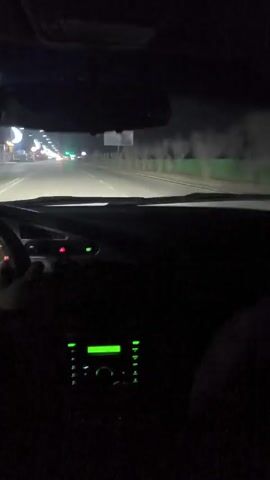 The video, shot from the driver's perspective inside a car, shows a night-time drive on a road illuminated by streetlights. The car's dashboard is visible, with a glowing speedometer, hazard lights, and other controls. Through the windshield, one can see a wide road with multiple lanes and sparse traffic. Streetlights, some shaped like crescent moons, line the road, along with traffic lights that are mostly green. The driver’s hands are visible on the steering wheel. In the audio, the beginning of a song plays, then someone yells loudly and laughs.