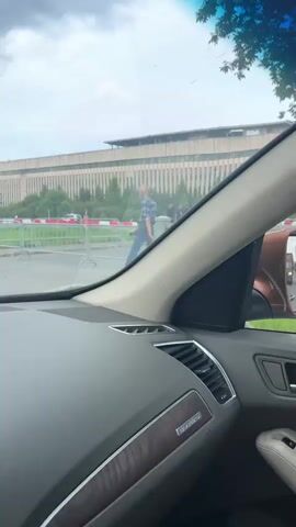 The video is shot from inside a car, looking out through the windshield. The car is moving, and the scene outside changes slightly throughout the short clip. In the background, there is a large, light-colored building with many windows, likely an airport terminal or a similar large public building. In front of the building, there are trees and a road or paved area. There are also some temporary red and white barriers. In the foreground, the dashboard of the car is visible, with a black and tan interior.

At the beginning of the video, there is a red and white barrier in the middle ground. Further back, there are trees and the large building.

As the video progresses, the view shifts. At 00:01, a person in blue shorts and a dark shirt is standing near a fallen bicycle.

By 00:02, the car has moved, and more trees and the building are visible. There are also people in the distance, some wearing high-visibility vests.

At 00:03, the view continues to change, showing more of the building and the surrounding area with trees and people.

At 00:04, the car passes a traffic sign showing a pedestrian crossing. There are also fire trucks visible in the distance, along with other vehicles and people.

At 00:05, the video ends, with the same general scene of the building, trees, and barriers visible. The sky is overcast.
