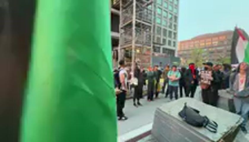 The video captures an outdoor daytime gathering or protest in an urban setting, likely a public square in Hoboken, United States. Approximately 20-30 individuals, mostly adults, are assembled in front of modern multi-story buildings, some featuring industrial-style metal frameworks. The sky is bright, indicating clear weather.

A large green flag partially obscures the foreground of the shot. Behind it, a person with a red, white, and black patterned keffiyeh-style headscarf is partially visible. To the right in the foreground, a rectangular stone or concrete block is present, with a black bag or item resting on it.

The central activity involves a speaker addressing the assembled crowd. This individual stands in the middle of the group, facing them, and is actively speaking. The crowd appears attentive and engaged. Several attendees are dressed casually, and some wear keffiyehs or other head coverings. Although difficult to discern clearly, some participants in the background appear to be holding signs.

At least one Palestinian flag (displaying red, black, white, and green) is clearly visible in the background, held by a participant on the right side of the crowd. During the video, a chant of "F*** you, Google!" is heard. The speaker then explicitly states, "And I'd like to call for all of us to end our relationship with Google too." This confirms the protest's target and nature, indicating an activist event directed at Google. The overall atmosphere suggests a unified demonstration of protest and advocacy.