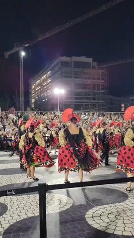 The video shows a nighttime outdoor performance with dancers in colorful costumes. The dancers are wearing red and black polka dot dresses with gold accents and some are holding fans. They are performing in front of a crowd of spectators. In the background, there is a large building under construction with cranes. The ground is paved with a patterned surface.