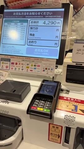 The video shows a person's hands interacting with a self-checkout kiosk in what appears to be a retail or restaurant setting. The kiosk has a touchscreen display with Japanese text. In the foreground, a hand is holding Japanese yen banknotes. The touchscreen displays payment options, including credit card, QR code, IC card, and cash. The person's finger is touching the screen, likely selecting a payment method. A payment terminal is also visible in the foreground. The background shows a display of sushi. The overall scene suggests a transaction is taking place at a point of sale.