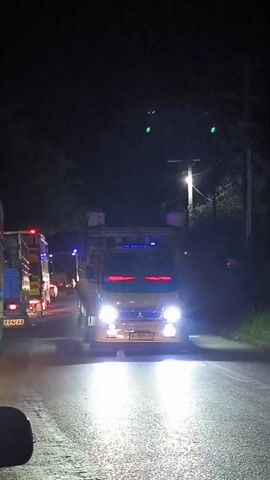 A light yellow/beige Mitsubishi truck drives on a road at night in Meuse, Indonesia. The truck approaches the camera, showcasing numerous bright lights: intense white headlights, red lights along the top of the windshield, blue lights beneath the windshield, and blue and green lights on the roof. A faint "Mitsubishi" logo is visible on the front grille. The license plate "BL 8560 AS" is clearly displayed on the front bumper. As the truck passes the camera, red side marker lights along its body become visible. The surroundings are dark with trees and what appear to be power lines or poles, indicating a nighttime setting in a rural or semi-urban area. The truck moves at a moderate speed, quickly passing by the viewer. The background becomes blurry after the truck passes.