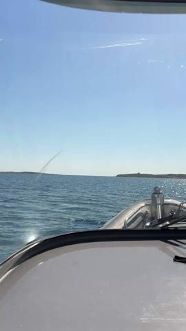 From inside a boat, a first-person perspective shot shows a clear blue ocean under a bright sky. Two small landmasses are visible in the distance on the horizon. The boat is moving, leaving small ripples on the water's surface. The camera pans slightly, revealing the boat's windshield and some equipment. A green "OPEN" sticker is visible on the side of the windshield.