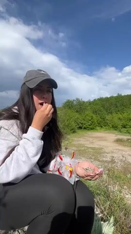 A woman in a baseball cap and a gray sweater sits eating sunflower seeds outside on a grassy, rocky patch. In the background, there are green trees and a cloudy, blue sky. The camera pans up, showing a panoramic shot of a mountain covered with trees and more cloudy, blue sky. The woman leans over to adjust her hair.