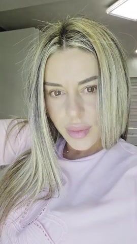 The video is a close-up selfie of a woman with blonde and brown hair, wearing a pink shirt. She is looking directly at the camera and speaking. The background is blurred but appears to be an indoor setting with some shelving or cabinets.