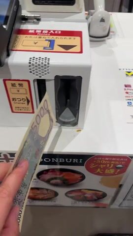 A hand inserts a 5000 yen banknote into a white cash-accepting machine. The banknote is fed into a slot, and a blue light appears within the slot. The scene then shifts to a touchscreen display showing a payment confirmation. The screen displays a total amount of 4,290 yen, the amount paid as 5,000 yen, and the change due as 710 yen. Payment options including various digital payment methods and credit cards are visible on the screen. A handheld payment terminal is also present. The machine appears to be a self-checkout or payment kiosk in a retail or food service environment.
