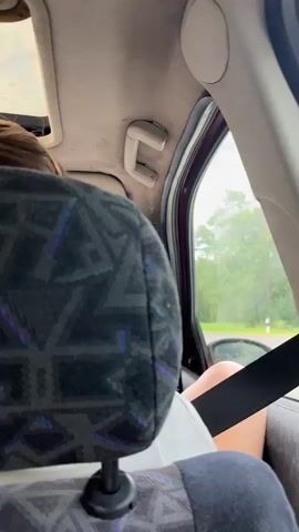 This video appears to be filmed from the back seat of a car, looking forward. The primary subjects are two people: a woman in the front passenger seat with her hair in a bun, and a man driving the car, wearing sunglasses and a light-colored t-shirt. The interior of the car is visible, including the headrests of the front seats, the dashboard with a rearview mirror, and a sunroof. Outside the car, a rural landscape with trees and greenery is visible, suggesting a daytime setting, likely overcast or rainy given the muted light.

The activity is a car journey. The man is actively driving, his hands on the steering wheel. The woman's posture suggests she might be looking out the window or resting. There are no discernible emotions conveyed in the brief clips, beyond a sense of casual travel.

Notable details include the patterned fabric of the car's headrests, the "smiley face" air freshener hanging from the rearview mirror, and the dashboard instruments. The location context provided indicates the journey is taking place in the Hartsjevichy Village Council area of Belarus. There is no visible text within the video frames themselves.