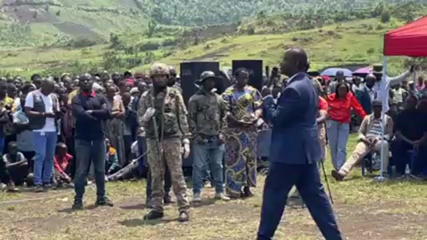 The video shows a man in a blue suit addressing a large crowd of people. He's holding a microphone and appears to be giving a speech. Behind him and to his side are several armed men in military camouflage. The crowd consists of people of various ages and genders, some standing and some sitting. There is a red tent visible in the background, as well as a backdrop of a green, hilly landscape. There are cameras and camera operators present, suggesting the event is being recorded. The setting appears to be an open, possibly rural area.