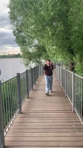 A woman walks towards the camera on a wooden boardwalk next to a body of water. The boardwalk has a metal railing on the left side. Green trees are visible on the right side of the boardwalk. The sky is cloudy. The woman is wearing a brown zip-up jacket over a white turtleneck and light blue pants. She has her hands in her pockets and is smiling.