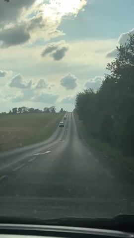 The video captures a point-of-view shot from a vehicle driving on a paved road that curves to the right and ascends a hill. The road is lined with trees on the right and a grassy field on the left. The sky is partly cloudy with the sun visible, casting shadows and creating lens flare. Several cars are ahead on the road, also heading uphill. The overall scene depicts a scenic drive through a rural landscape under a dynamic sky.