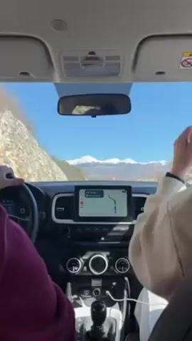 Here's a description of the video:

The video shows the interior of a car driving on a road with a mountain range in the background. The mountains are snow-capped. The car's dashboard and navigation screen are visible. The driver, wearing a purple sweatshirt, can be seen holding the steering wheel. A passenger, wearing a beige jacket, is also visible. The weather is sunny. The navigation screen shows a map with a route and distances. The video is shot from the perspective of the driver's seat. Music is playing in the background.