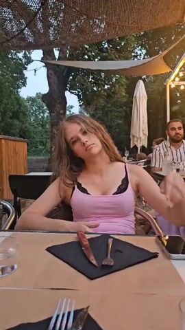 A young woman with long, light brown hair sits at a table in an outdoor dining area. She is wearing a pink tank top with black lace trim. She has her right hand raised to her head and is touching her hair. She looks off to the side with a neutral expression. In front of her on the table are a knife, a fork, a black napkin, and a glass of water. Behind her, a man with dark hair and a beard is visible at another table. The background shows trees and a canopy overhead with string lights.