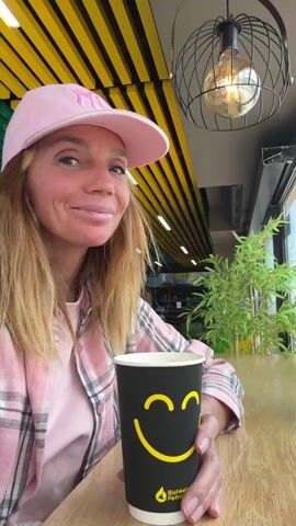 A woman in a pink plaid shirt and pink baseball cap holds a black paper cup with a yellow smiley face on it. The cup says “Bishkek Petrol” at the bottom. She smiles at the camera and raises the cup. She appears to be sitting at a table in a cafe.  Behind her is a yellow and black striped ceiling, a hanging light fixture, and a window with green plants.