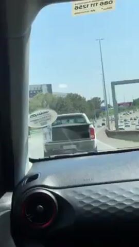 The video shows a view from inside a car driving on a busy highway. The car is in the right lane, following a silver pickup truck. The sky is blue, and there are several light poles lining the side of the highway. Trees are visible on the side of the road, as is a tall building in the distance. The dashboard of the car in the foreground is black. The interior of the car and the windshield obstruct some of the view.