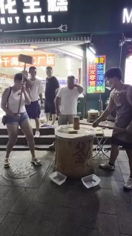 A group of people are gathered around a large, cylindrical, wooden object on a sidewalk at night. A man wearing an apron stands to the right of the object, holding a large mallet and striking the object. A woman in a white tank top and denim shorts stands to the left of the object, also holding a mallet and appears to be waiting her turn or has just hit the object. Other individuals, including a man holding a baby, are standing behind the woman, observing the activity. The wooden object has Chinese characters inscribed on it and appears to be part of a street performance or vendor stall. The environment is lit by neon signs and store lights.