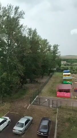 The video shows an aerial view of a residential area with trees, a playground, and cars parked. The weather appears overcast, possibly rainy, as evidenced by the texture on the window pane in the foreground which suggests rain streaks. The playground is colorful with various structures like slides, swings, and small playhouses. There are three cars visible in the parking lot, two sedans and a hatchback. A fence separates the parking area from the playground and a row of trees. In the background, there are more trees and some buildings, suggesting a rural or semi-rural setting. The sky is cloudy, with a circular, slightly hazy light spot that might be an artifact on the camera lens or window, or possibly a distant light source. The overall impression is of a quiet, somewhat damp day.