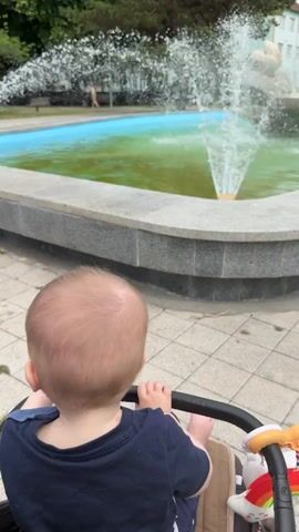 A baby is seated in a stroller facing away from the camera, looking at a fountain with turquoise water. The fountain is spraying water upwards, creating a misty effect. The fountain is surrounded by a stone border and is located in an outdoor area with trees and buildings in the background. The ground around the fountain is paved with square tiles. The sky is overcast.