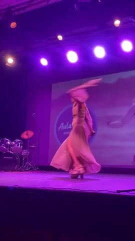 A woman with blonde hair, wearing a light-colored, flowing dress with a slit on the side and a scarf that trails behind her, stands on a stage. She is holding a microphone in her right hand and appears to be mid-performance, with her body turned to her left and her dress fanned out around her. A drum set is visible to the left of the stage, and a large screen behind her displays a logo that reads "Aula de Visión SONG CONTEST" with a heart graphic. The stage is lit with purple spotlights.