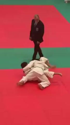 Two judokas in white uniforms with green belts are grappling on a red and green judo mat. One judoka is on top, attempting to control the other. A referee in a black suit is standing nearby, observing the match. The referee extends his arm toward the judokas, signaling something.