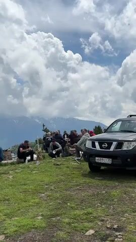 This video shows a group of people gathered in a mountainous, outdoor setting. They appear to be having a picnic or a casual social gathering. A black SUV is parked on the right side of the frame. The background consists of lush green hills and distant snow-capped mountains under a cloudy sky. Some individuals are sitting on the grass, while others are standing and interacting. The overall atmosphere suggests a relaxed, informal event in a scenic natural landscape.