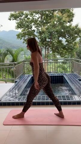 A woman in a leopard print jumpsuit is performing a yoga pose on a pink mat. She stands with her legs wide apart, one foot angled slightly inward and the other pointing straight ahead. Her arms are clasped behind her back. She then bends forward from her waist, extending her left arm towards the ceiling and her right arm towards the floor. The background features a balcony with a railing, a swimming pool, lush green trees, and mountains in the distance.