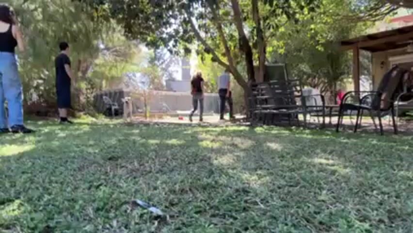 In a sunlit backyard, four people, two women and two men, are visible alongside a large, light-colored dog. The dog is playing, retrieving a stick. One woman throws the stick. The yard has a grassy lawn with patches of bare earth, surrounded by trees and bushes. In the background, there's a fence and what appears to be another building. Outdoor chairs and tables are clustered near a tree, and part of a covered patio is visible on the right side of the frame.