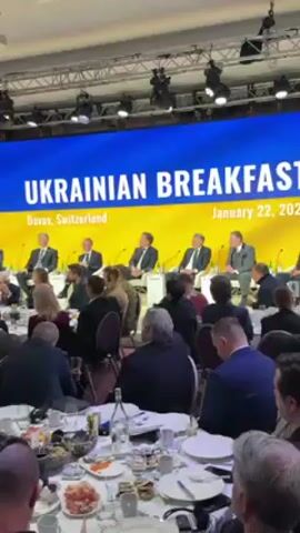 The video depicts a gathering at what is described as a "Ukrainian Breakfast" held in Davos, Switzerland on January 22, 2026. Attendees are seated around large tables with plates, glasses, and bottles of water. A panel of individuals is seated on a stage, presumably speaking at the event. Most attendees are facing the panel, listening. The environment appears to be a well-lit conference room with a large screen displaying the event title. Individuals are generally dressed in professional attire.