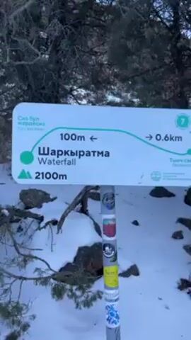 The video begins with a shot of a sign in a snowy, mountainous area. The sign reads "Сіз бұл жердесіз You are here" in both an unfamiliar script and English. It also indicates that "Шаркыратма Waterfall" is 100 meters to the left, and "Тыныш сай Backwater" is 0.6 kilometers to the right. The elevation is listed as 2100 meters. The camera pans up to show the snowy landscape, revealing sparse trees and a bright, sunny sky. A woman appears on a snowy path, wearing a green jacket, a black hat, and a backpack. She is looking at the camera and smiling, with snow-capped mountains in the background.