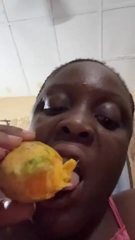 A close-up video shows a Black woman eating a mango. She holds the mango in her hand, taking bites from it. The mango is yellow with hints of green on the skin, and the flesh is orange. The background features a white tiled ceiling and a partially visible wall with a light color. The woman is wearing a pink top. Her face is close to the camera, showing her eyes, nose, and mouth.