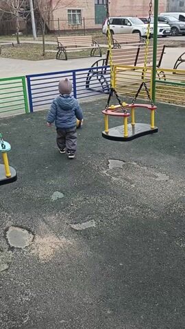 The video shows a toddler wearing a gray hat and a blue jacket walking around a playground with a volleyball. The playground has a soft, dark green surface and is equipped with several swings with yellow, green, and red accents. Colorful, low fences surround the play area. In the background, there are residential buildings, parked cars, benches, and a few adults sitting. The toddler moves around the swings, occasionally kicking the volleyball.