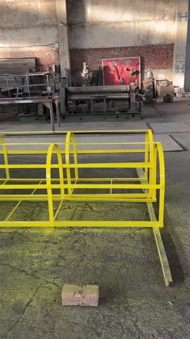 This media file shows a bright yellow metal structure, possibly a ladder or railing, lying horizontally on the floor of a workshop or factory. The structure is made of yellow metal bars and features arched supports. In the background, there are various pieces of industrial machinery, including what appears to be a large metal rolling machine and other equipment. The setting is a workshop with concrete floors, brick walls, and large windows, suggesting an indoor industrial environment. There is some debris on the floor, including a brick in the foreground. The lighting suggests it could be daytime, with sunlight streaming in through the windows. No people are visible in the frame. No visible text is present. The overall impression is of a functional, industrial space where metal fabrication or repair work is taking place.