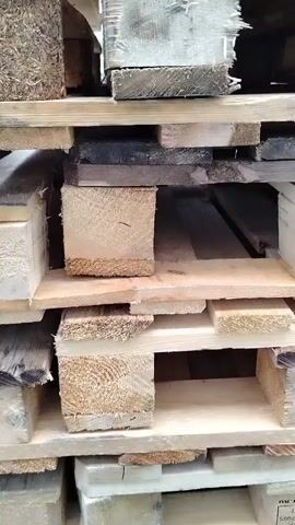 The video shows a close-up, vertical shot of stacked wooden pallets. The pallets are made of rough, unfinished lumber of various sizes and colors, some appearing old and weathered, others newer. The wood grain and texture are clearly visible. The pallets are assembled in a typical pallet structure with horizontal planks supported by vertical blocks. The camera angle is static, and the lighting appears to be natural, casting soft shadows. The background is out of focus, suggesting the pallets are in a storage area. No people or actions are visible; it is a still life of industrial materials.