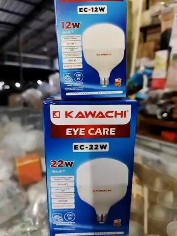 The video shows two boxes of Kawachi Eye Care LED light bulbs stacked on top of each other. The box on top is for a 12W bulb, and the box on the bottom is for a 22W bulb. The boxes are blue and red with white text. A light-colored tiled roof and a blurry background are visible.