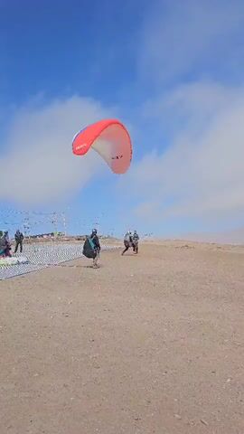 The video shows a person preparing for paragliding. The individual, dressed in dark clothing and a helmet, is standing on a sandy surface. Above them, a red, white, and blue paragliding wing labeled "TONIC" is inflated. Two other people are nearby. The sky is blue with scattered clouds, and in the distance, there appears to be a city or settlement. The wind is strong enough to inflate and lift the paragliding wing. The person seems to be adjusting equipment and getting ready for takeoff.