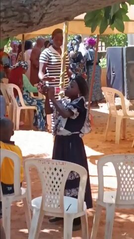 The scene appears to be an outdoor gathering, possibly a celebration, held under a large white tent. Numerous people are visible, some seated on white plastic chairs and others standing. A woman in a long black and white striped dress stands near the edge of the tent. A young girl in a dark dress with a patterned top stands in the foreground, appearing to be filming or taking pictures with a mobile device. The environment seems bright and sunny, with trees visible in the background, creating a dappled lighting effect.