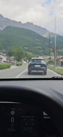 The video begins with a view from inside a car, looking forward through the windshield. A dark-colored SUV is visible driving ahead on a road. In the background, there are buildings, trees, and steep, majestic mountains. The camera then pans slightly to the right, showing the side of the car and a partial view of the passenger window. The scene outside continues to reveal a road with buildings and trees, with the mountains forming a dramatic backdrop. The perspective shifts again to show more of the side of the car and the exterior landscape. The video continues to capture the view from the moving vehicle, showcasing a picturesque mountain village with traditional houses, green fields, a wooden structure, and lush trees, all set against the backdrop of imposing mountains under a partly cloudy sky.