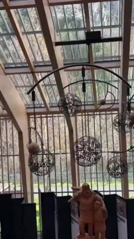 The video shows the interior of a building, possibly a museum or exhibition hall. The structure has a high glass ceiling supported by a wooden framework, allowing natural light to flood the space. In the foreground, there's a wooden sculpture of two figures, an adult and a child. Behind the sculpture, there are black panels or dividers, presumably part of the exhibit. Hanging from the ceiling is a decorative metal structure consisting of curved rods and several spherical, cage-like elements. The spheres have some writing on them. Through the glass ceiling and windows, trees and foliage are visible. The overall impression is that of a well-lit, curated space showcasing art and possibly historical artifacts.