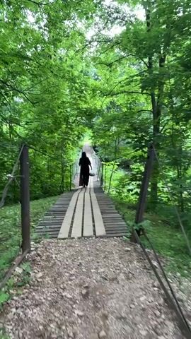 A person walks across a wooden footbridge in a forest. The bridge is made of weathered planks and appears to be suspended. The person is silhouetted against the greenery and is walking away from the camera. The surrounding environment is lush and dense with trees, with sunlight filtering through the leaves. The ground leading up to the bridge is covered in gravel and small stones. The overall atmosphere is peaceful and natural.