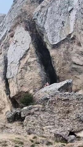 A person in a pink shirt is sitting on a large rock formation at the base of a tall, rugged mountain range. The person is looking towards the mountain face. The mountain is composed of light brown and grey rock, with a deep crevice in the center. There is dry grass and some rubble at the base of the mountain. The sky is overcast.