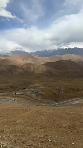 The video shows a high-angle, stationary view of a mountainous landscape under a cloudy sky. The terrain is primarily arid and brown, with rolling hills stretching into the distance. In the valley below, a winding road with multiple switchbacks is visible, with a few vehicles traveling along it. Distant snow-capped mountains are visible on the horizon.  The sky is overcast with large, grey and white clouds.