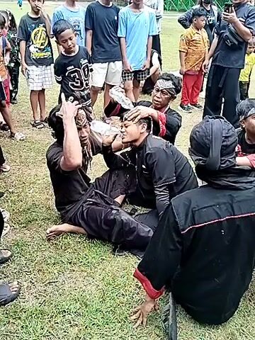 The video shows a group of people in an outdoor area, seemingly a park or grassy field. Most are young men and boys, dressed in casual attire and what appears to be traditional Indonesian clothing, particularly black outfits with headbands. One individual, sitting on the ground, is having water poured over their head by another person, also seated, while others assist and observe. There is a festive or celebratory atmosphere with smiles and laughter visible. A banner in the background indicates an event or gathering, though the details on the banner are difficult to discern clearly. The environment is grassy with trees visible in the background.