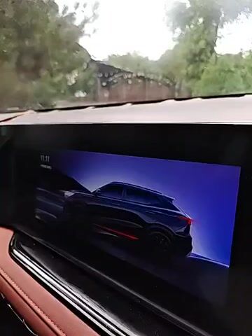 The video shows the interior of a car. A large touchscreen display in the center of the dashboard shows an image of a dark blue SUV with red accents. The steering wheel is visible in the foreground. The windshield is visible in the background and appears to be wet, suggesting it is raining. The car's interior is a dark color with red stitching on the seats and dashboard. The overall impression is a modern and luxurious vehicle.