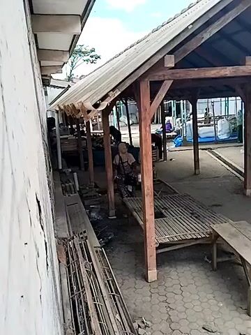 The video shows a person wearing a head covering and an apron standing next to a bamboo bench in an open-air shelter. The person is holding a white bucket and appears to be engaged in some activity, possibly painting or cleaning. The shelter has a corrugated metal roof supported by wooden beams. In the background, other structures and people are visible, along with stacks of materials. The ground is paved with interlocking concrete blocks. The overall environment suggests an outdoor market or workshop area.