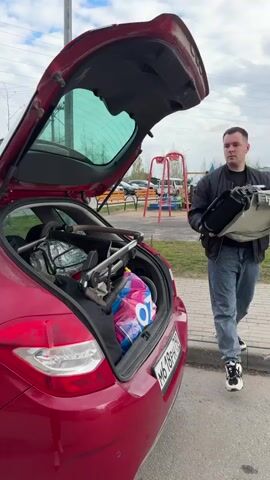 An adult male is seen loading a folded baby stroller into the open trunk of a red hatchback car. The car, possibly a Citroën C4, is parked outdoors on an asphalt surface. The man, dressed in a black jacket, blue jeans, and white and black sneakers, carefully maneuvers a folded beige/khaki baby stroller into the trunk. The trunk already contains several colorful plastic bags and other items.

The daytime scene is under an overcast sky. In the background, a utility pole with overhead wires, bare-branched trees, and a red metal playground structure are visible, indicating a residential or suburban setting. The car's Russian license plate, 'M 618 PH 196', is clearly visible on the rear. The man appears focused as he secures the stroller in the limited trunk space.